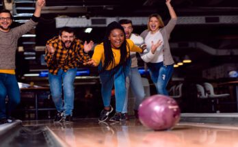 A group of friends smile, yell, and cheer as they watch a mauve bowling ball roll down a bowling lane.