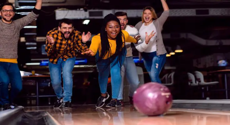 A group of friends smile, yell, and cheer as they watch a mauve bowling ball roll down a bowling lane.