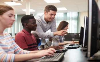 A teacher leans over a young high school student's shoulder to point at a desktop monitor in a computer classroom.