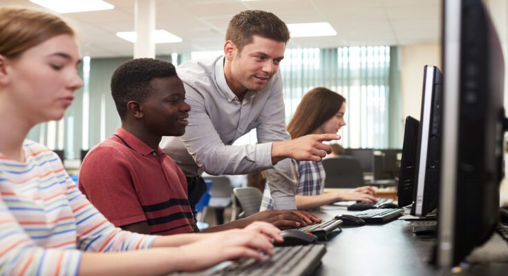 A teacher leans over a young high school student's shoulder to point at a desktop monitor in a computer classroom.