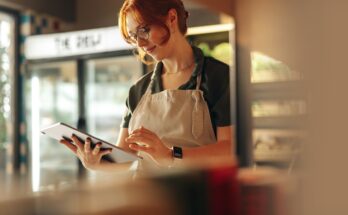A woman with red hair, wearing a green uniform and glasses, stands in a well-lit store using a tablet.