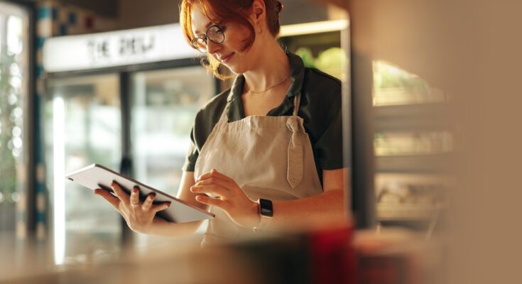 A woman with red hair, wearing a green uniform and glasses, stands in a well-lit store using a tablet.