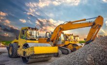 A drum roller is parked next to a crane and another drum roller. A pile of rock and gravel is in front of the equipment.
