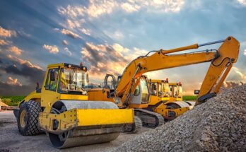 A drum roller is parked next to a crane and another drum roller. A pile of rock and gravel is in front of the equipment.