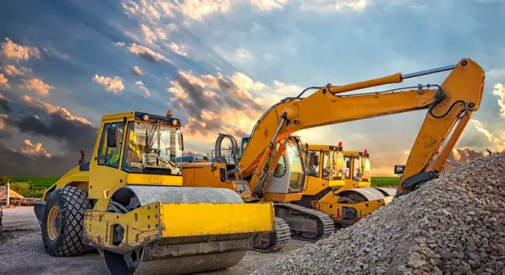 A drum roller is parked next to a crane and another drum roller. A pile of rock and gravel is in front of the equipment.