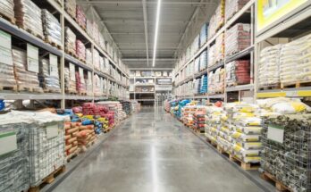 A warehouse aisle with shelves stocked with supplies, all placed on wooden pallets on both the floor and the shelving.