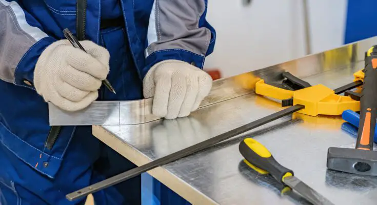 A metalworker marking a piece of metal with a marker. There are multiple measuring tools next to his workspace.