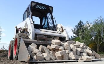 A white skid steer with red wheels has a bucket attachment full of stone bricks