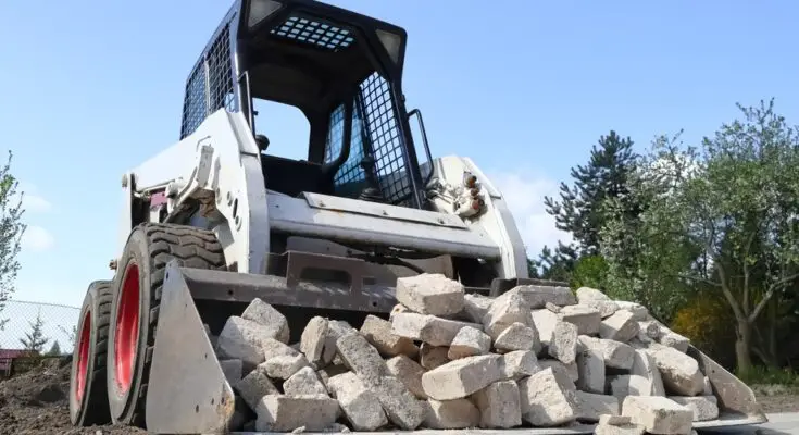 A white skid steer with red wheels has a bucket attachment full of stone bricks