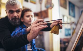 A father shows his son how to hold a paintbrush as they paint together on a canvas in an art studio.