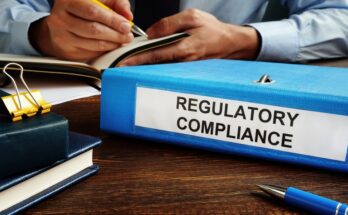 A binder labeled "Regulatory Compliance" sits on a businessperson's desk, next to two books and a pen.
