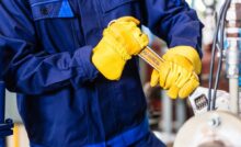 A mechanic wearing navy blue coveralls and yellow gloves holds a wrench and tightens a component on a machine.