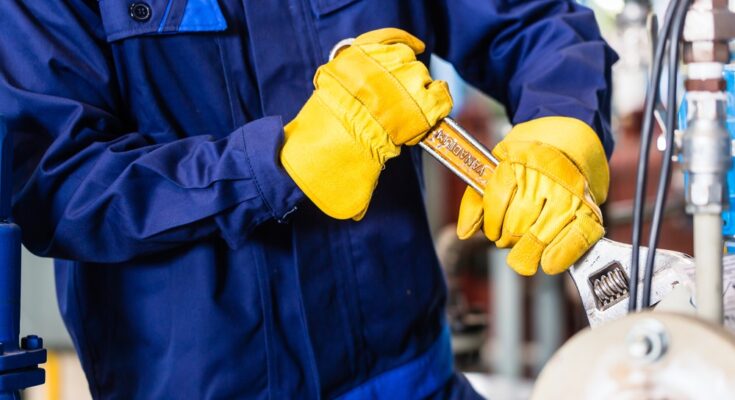 A mechanic wearing navy blue coveralls and yellow gloves holds a wrench and tightens a component on a machine.