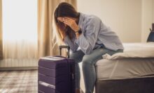 A woman sits on a hotel bed, her head in her hands, looking tired and frustrated. Her suitcase is in front of her.