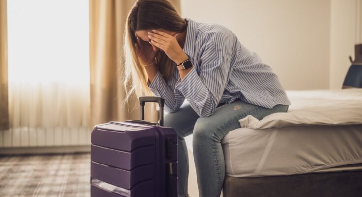 A woman sits on a hotel bed, her head in her hands, looking tired and frustrated. Her suitcase is in front of her.