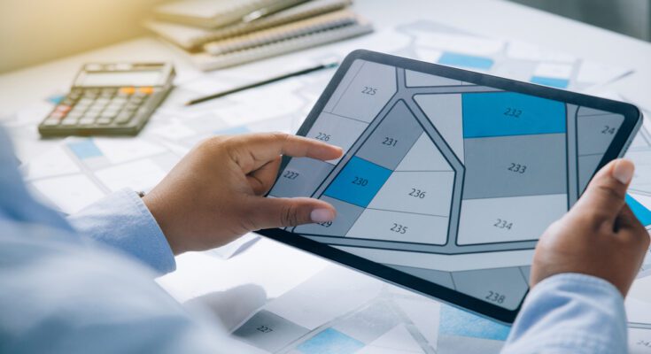 A man's point of view as he examines a map of land parcels on an electronic tablet, with some lots shaded in blue and grey.
