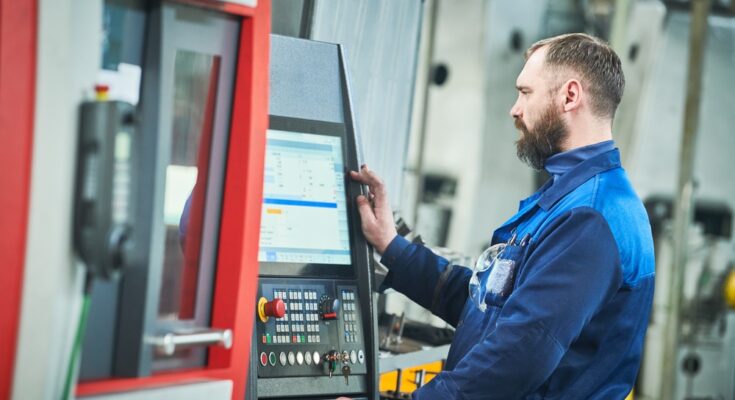 A CNC machine operator in a blue jumpsuit inspects a screen of data analytics attached to his CNC machine.