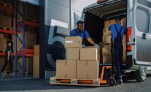 Two men stand outside a warehouse, stacking cardboard boxes of varying sizes into the back of a truck.