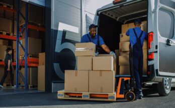 Two men stand outside a warehouse, stacking cardboard boxes of varying sizes into the back of a truck.