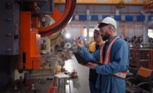 Two technicians in a facility look at a piece of machinery and discuss it. Both wear hard hats and carry laptops or tablets.