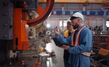 Two technicians in a facility look at a piece of machinery and discuss it. Both wear hard hats and carry laptops or tablets.