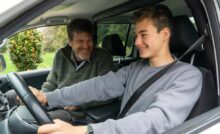 A father sits in the passenger seat of a car while his younger son sits in the driver's seat. Both are smiling.