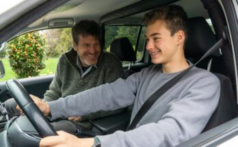 A father sits in the passenger seat of a car while his younger son sits in the driver's seat. Both are smiling.