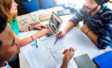 Three people sit at a table. One types on a computer while the other two point to details on a blueprint.