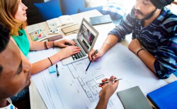 Three people sit at a table. One types on a computer while the other two point to details on a blueprint.