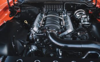 A close-up of an orange vehicle's open hood reveals a large engine with an intricate system of parts.