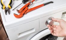 Pliers, a screwdriver, and a clamp sit on top of a front-load washer machine; someone’s hand is on the knob of the appliance.