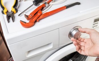 Pliers, a screwdriver, and a clamp sit on top of a front-load washer machine; someone’s hand is on the knob of the appliance.