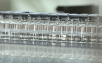 A row of small glass vials with grey lids that each contain a clear liquid on a reflective surface.