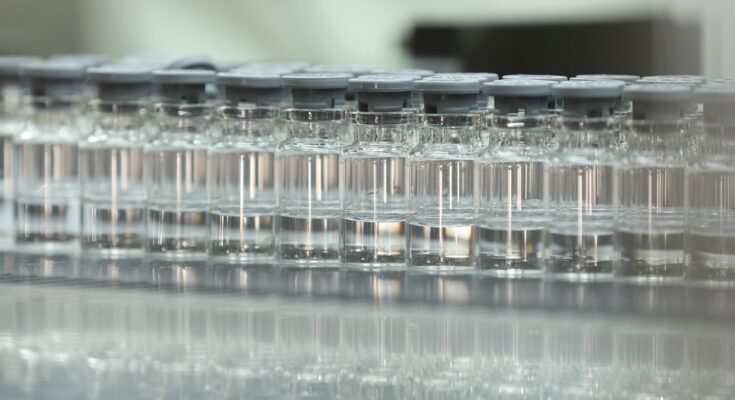 A row of small glass vials with grey lids that each contain a clear liquid on a reflective surface.