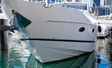 A close-up of the hull of a large white and black boat docked at a marina with tall buildings in the background.