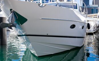 A close-up of the hull of a large white and black boat docked at a marina with tall buildings in the background.
