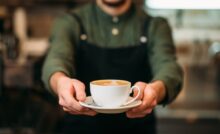 A person wearing a green shirt and black apron holds out a white cup of coffee on a small plate with both hands.