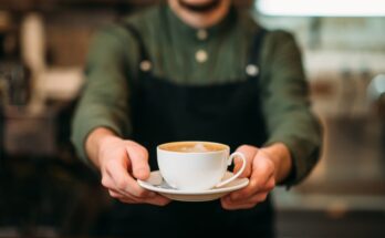 A person wearing a green shirt and black apron holds out a white cup of coffee on a small plate with both hands.