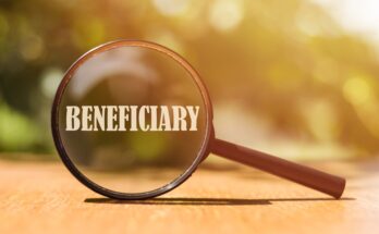 A brown handheld magnifying glass sits on a wooden surface, magnifying the cream-colored word "beneficiary."