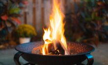 A close-up view shows a handful of logs on fire in a dark-colored fire pit. Landscaping appears blurry behind the pit.