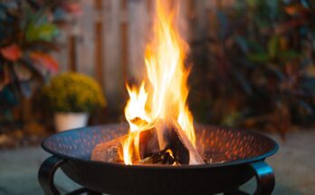 A close-up view shows a handful of logs on fire in a dark-colored fire pit. Landscaping appears blurry behind the pit.