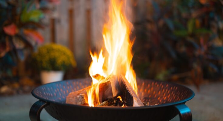A close-up view shows a handful of logs on fire in a dark-colored fire pit. Landscaping appears blurry behind the pit.