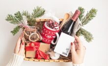 A basket is filled with a bottle of wine, a box with a red bow, a mug with a sweater pattern, and a few spruce leaves.