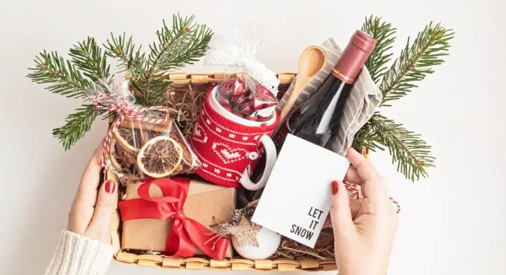 A basket is filled with a bottle of wine, a box with a red bow, a mug with a sweater pattern, and a few spruce leaves.