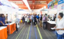 A blurred view of a trade show walkway that's lined with booths on both sides. People are walking and observing the booths.