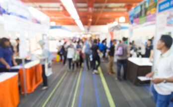 A blurred view of a trade show walkway that's lined with booths on both sides. People are walking and observing the booths.