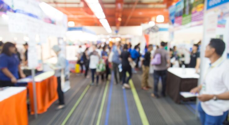 A blurred view of a trade show walkway that's lined with booths on both sides. People are walking and observing the booths.