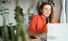A young woman with curly hair and glasses holds a pen to her face as she works from home on her laptop.