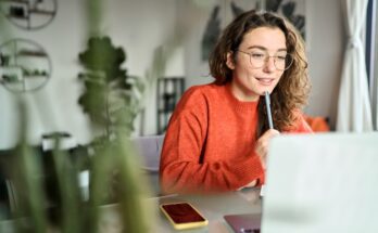 A young woman with curly hair and glasses holds a pen to her face as she works from home on her laptop.
