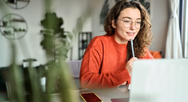 A young woman with curly hair and glasses holds a pen to her face as she works from home on her laptop.